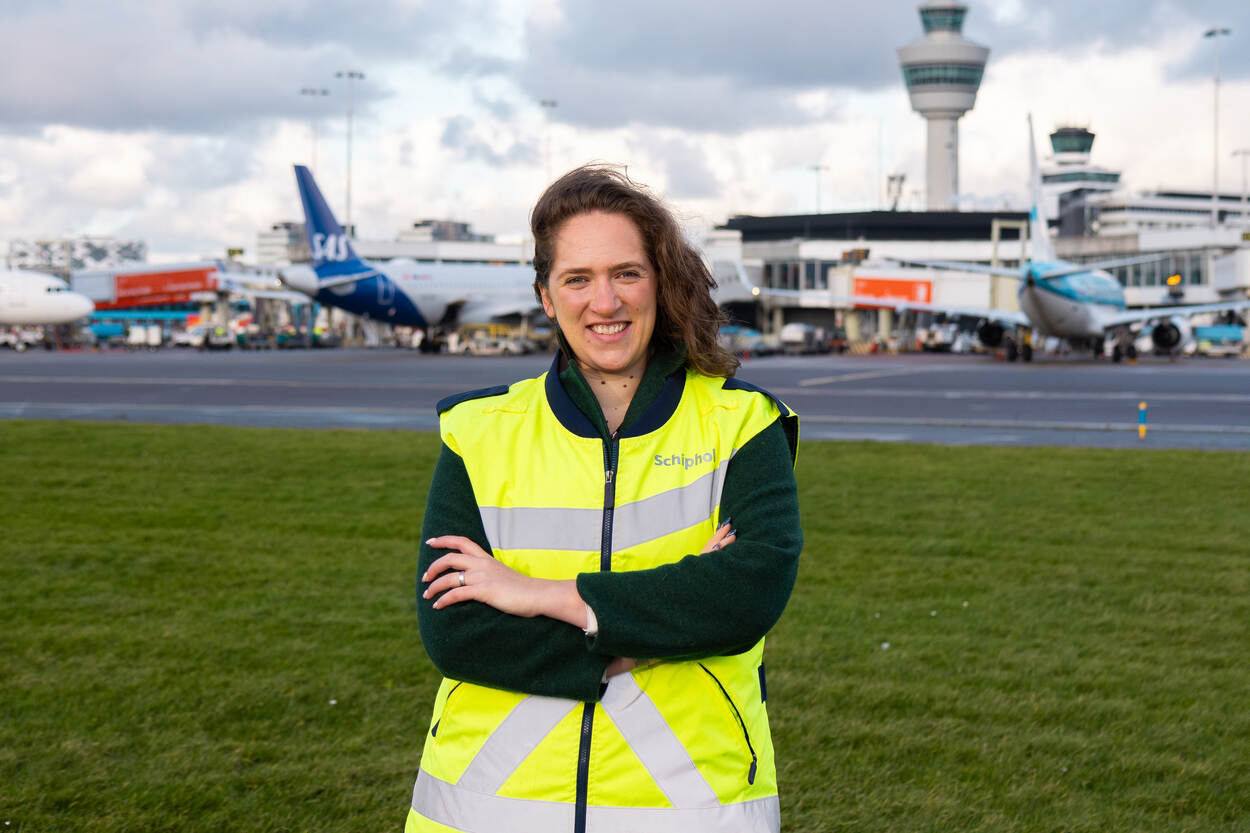 Anne 's-Gravenmade, Flow Manager Aircraft op Schiphol, staat met een veiligheidshesje buiten op de luchthaven, met op de achtergrond vliegtuigen aan de gates