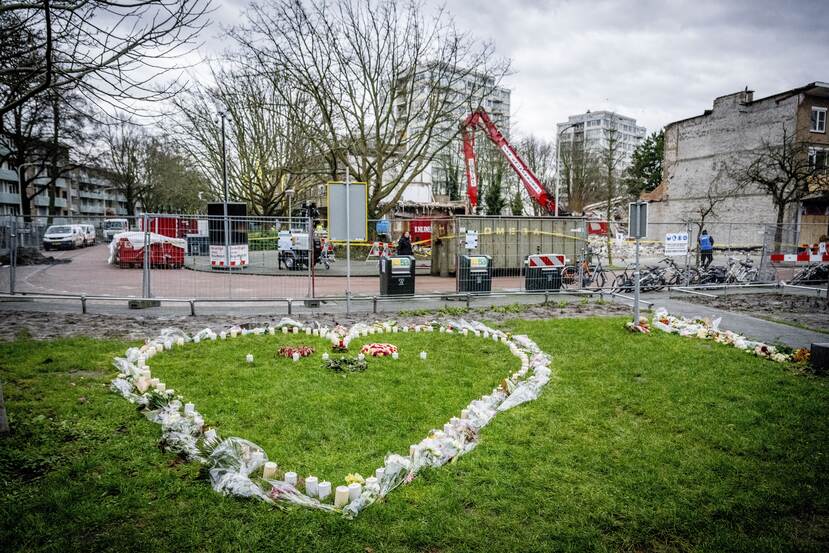 Een bloemenzee en kaarsjes in de vorm van een hart op een grasveld, met op de achtergrond graafmachines die de ravage opruimen na de explosie op de Tarwekamp