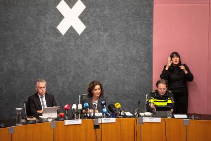 Een persconferentie in Amsterdam, met zittend in het midden de burgemeester Femke Halsema en aan haar zijden de officier van justitie en het hoofd van de Amsterdamse politie. Op de achtergrond een gebarentolk, die staat.